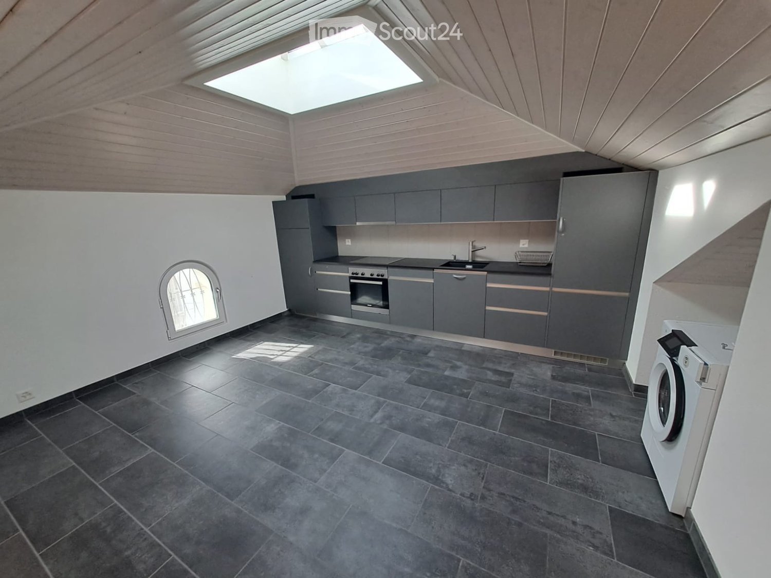 Modern kitchen with grey cabinets, dark tiled floor, skylight window, and built-in oven and sink.