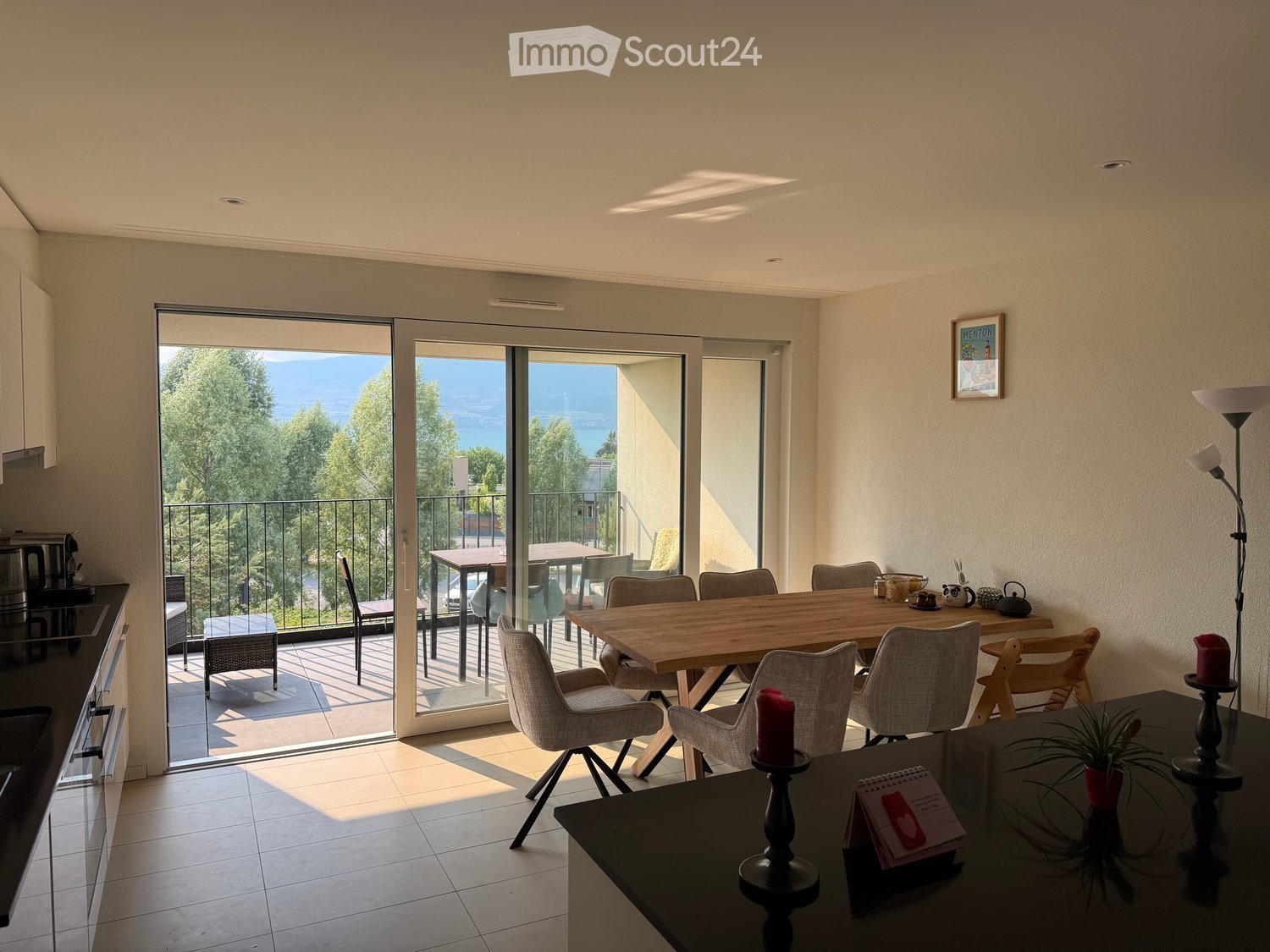 Bright, open-plan living and dining area with large sliding glass doors leading to a balcony with scenic views. The space includes a wooden dining table with chairs, a coffee table with a candle and plant, and a microwave on the counter.
