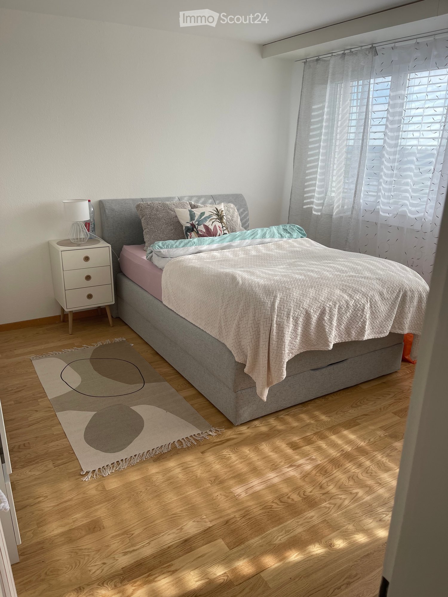 Modern bedroom with a grey bed, pink bedding, a nightstand with a lamp, and a decorative rug on a wooden floor.