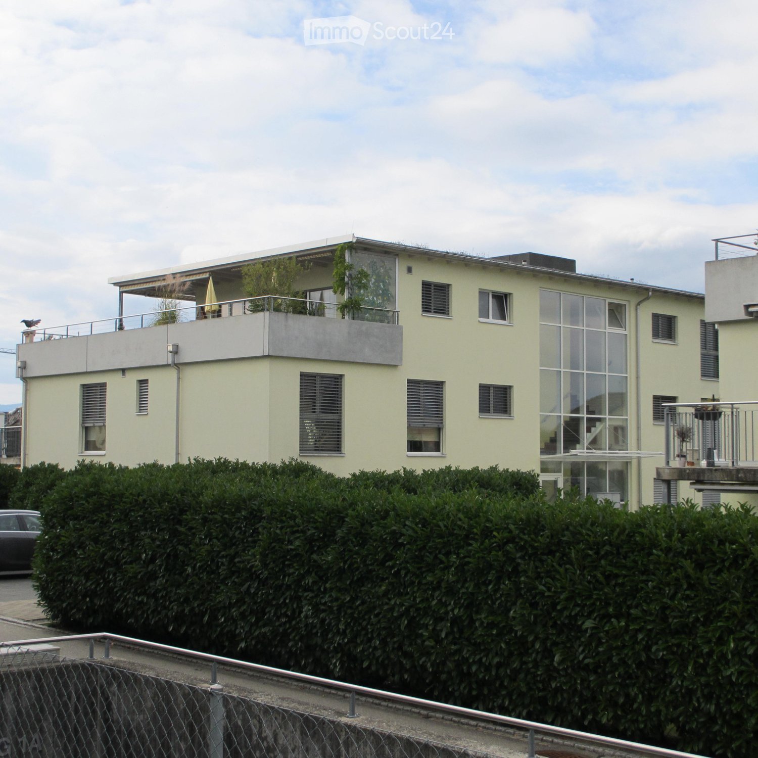 3-storey building, large glass windows, top balcony with metal railing, front garden with bushes