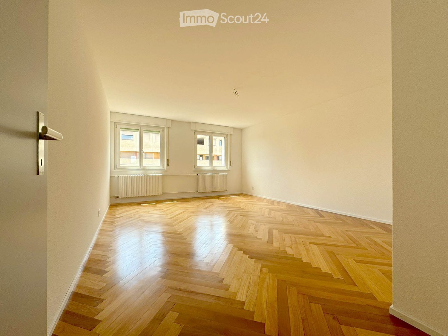 Empty room with wooden herringbone flooring, white walls, two windows, radiators, and a door handle visible.
