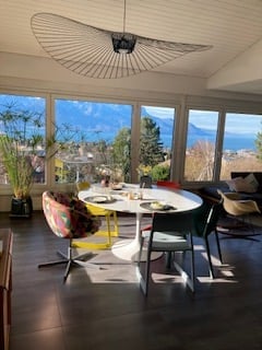 Bright, modern dining area with large windows offering a scenic mountain view. Features a round white dining table with colorful chairs, a potted plant, and a contemporary floor lamp.
