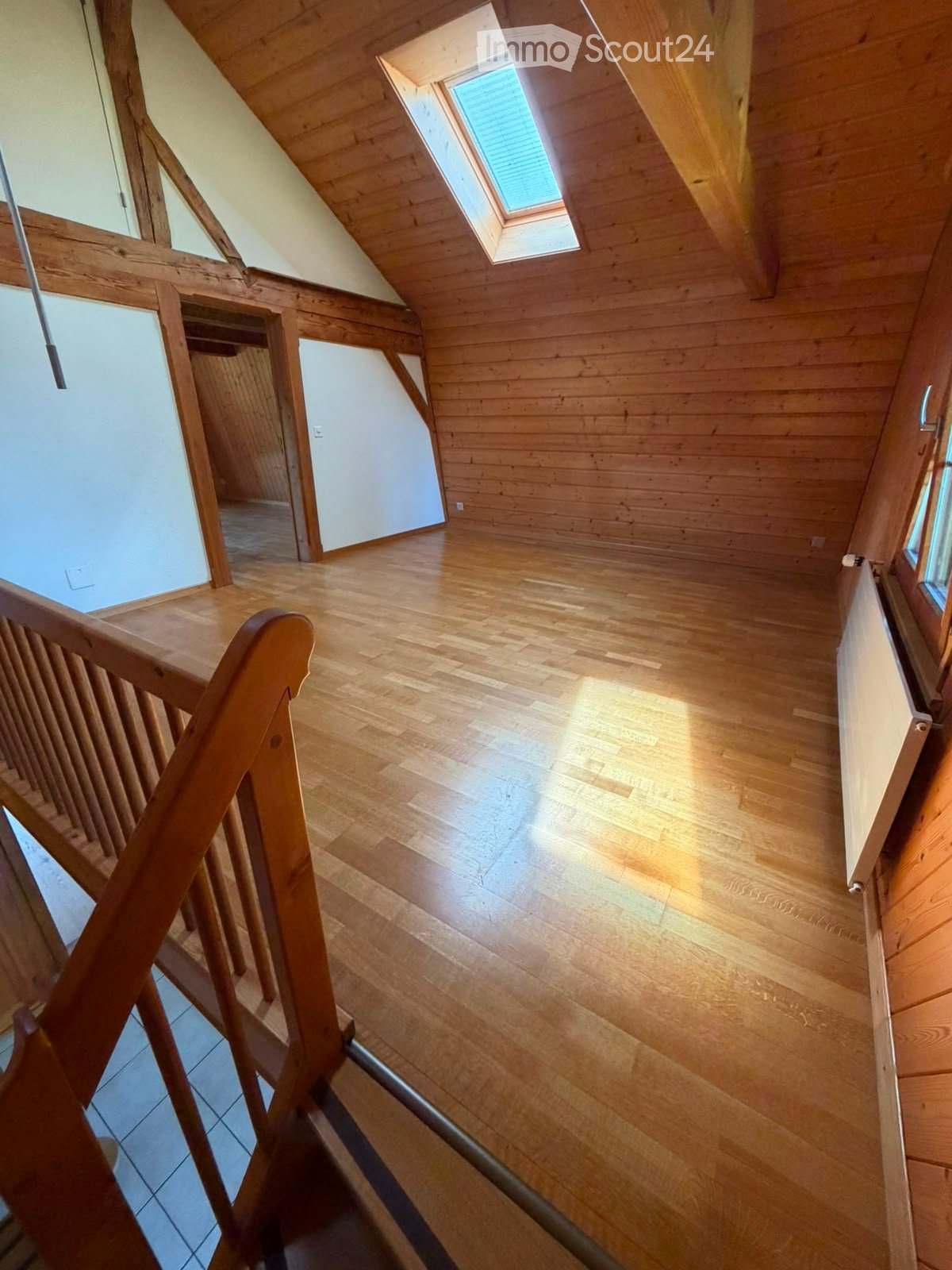 Bright, wooden-floored attic space with exposed wooden beams, skylight window, and wooden staircase leading downwards.