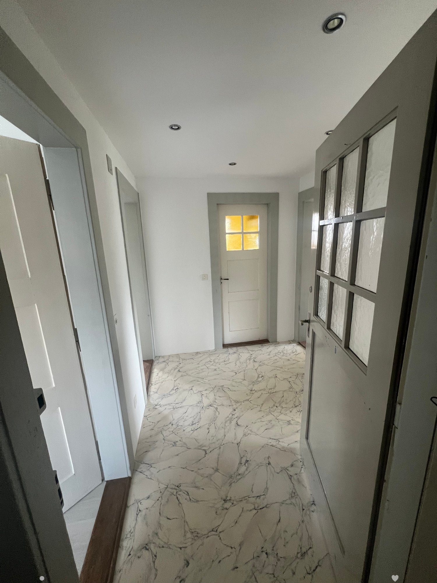 A long hallway with marble-patterned flooring, white walls, and several doors, including one with a frosted glass panel.