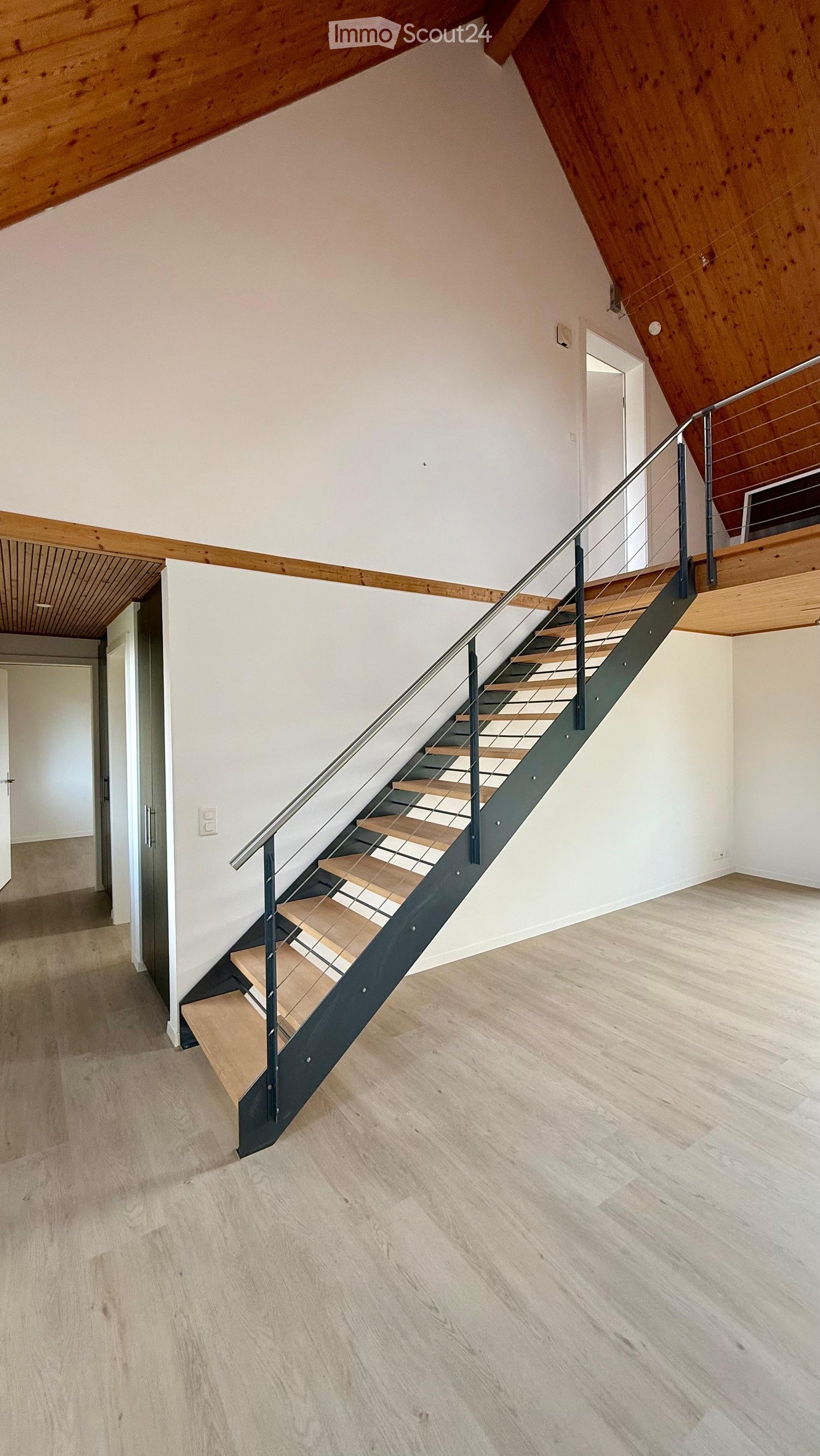 Modern hallway with wooden flooring, white walls, and a staircase featuring wooden steps and a metal railing. The ceiling has exposed wooden beams, and there are doors visible on either side.