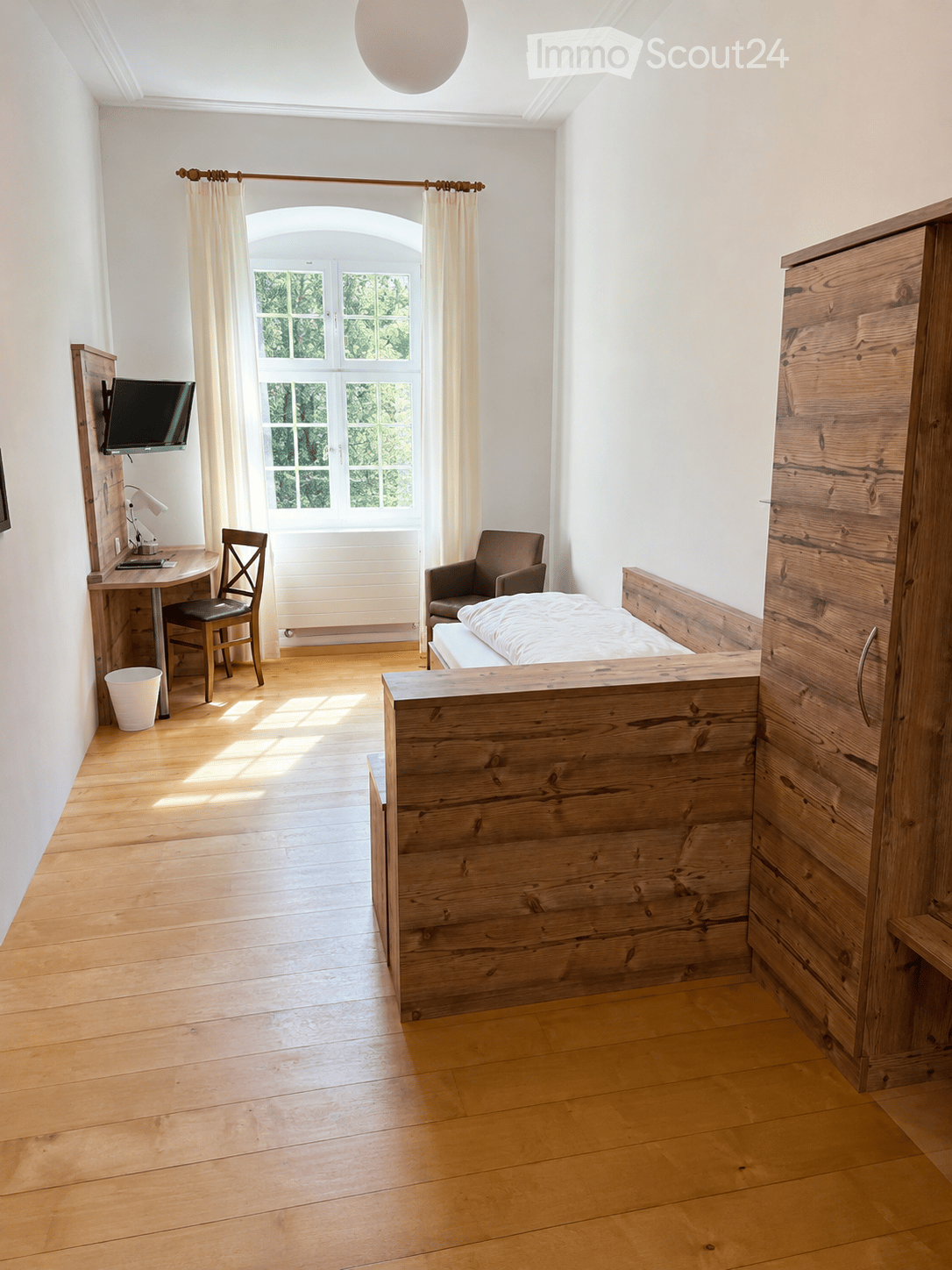 Cozy bedroom with wooden flooring, a bed with a wooden headboard, a chair, a desk with a screen, and a wardrobe.