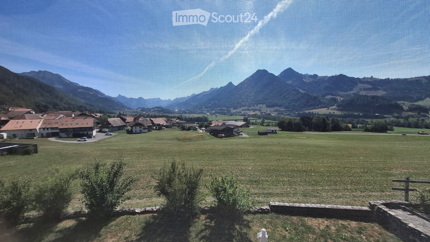 A panoramic view of a rural landscape with green fields, scattered houses, and distant mountains under a clear blue sky.