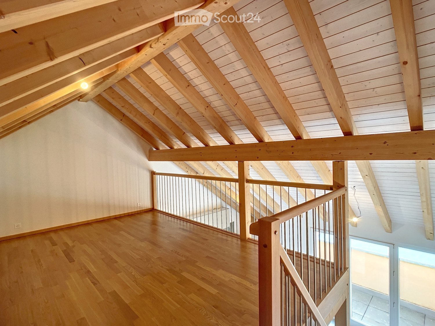 Open, airy hallway with exposed wooden beams and a modern metal railing leading to a staircase. The floor is made of polished hardwood, and the walls are painted in a light neutral tone.
