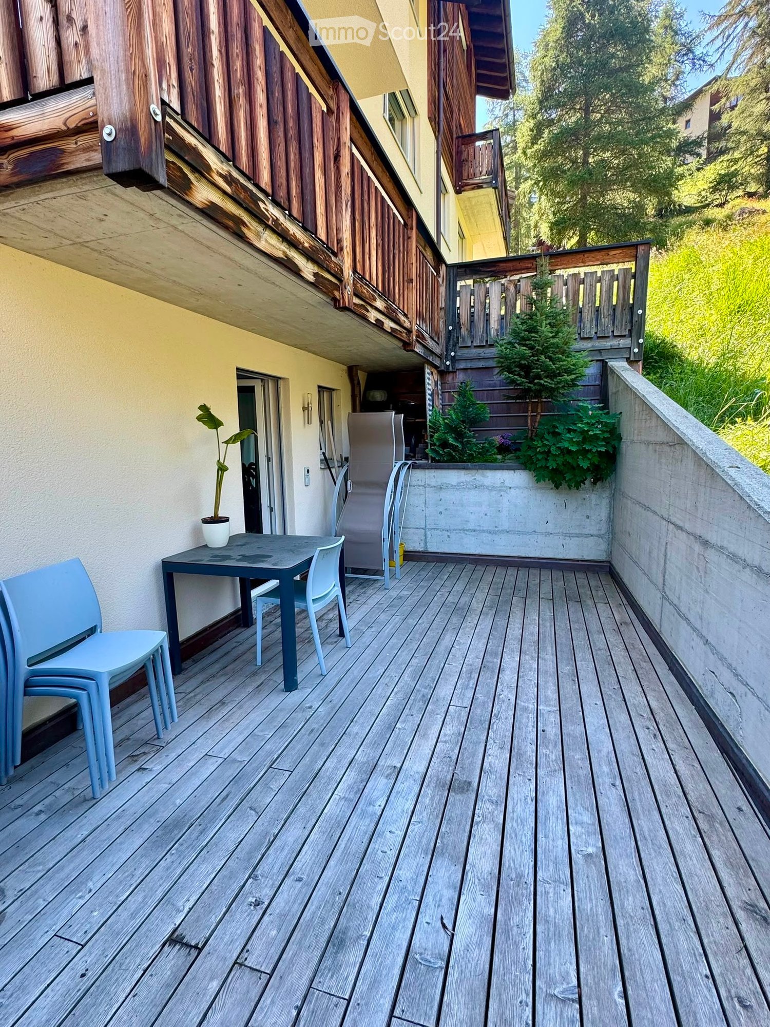 Wooden deck with a small table, chairs, and a potted plant, adjacent to a building with a staircase leading up.