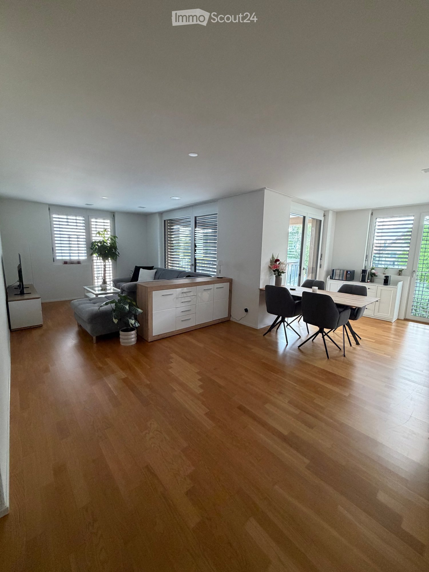 Open-plan living and dining area with wooden flooring, white walls, and large windows with blinds. Features a grey sofa, ottoman, coffee table, dining table with chairs, and a cabinet.