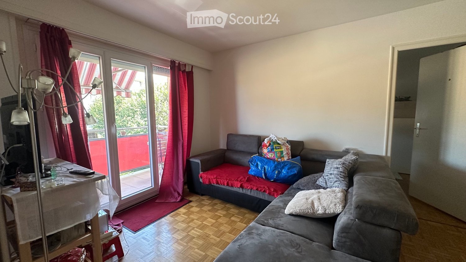 Living room with wooden flooring, large sliding glass doors leading to a balcony, grey sectional sofa with pillows, coffee table, floor lamp, and a small table with items on it.