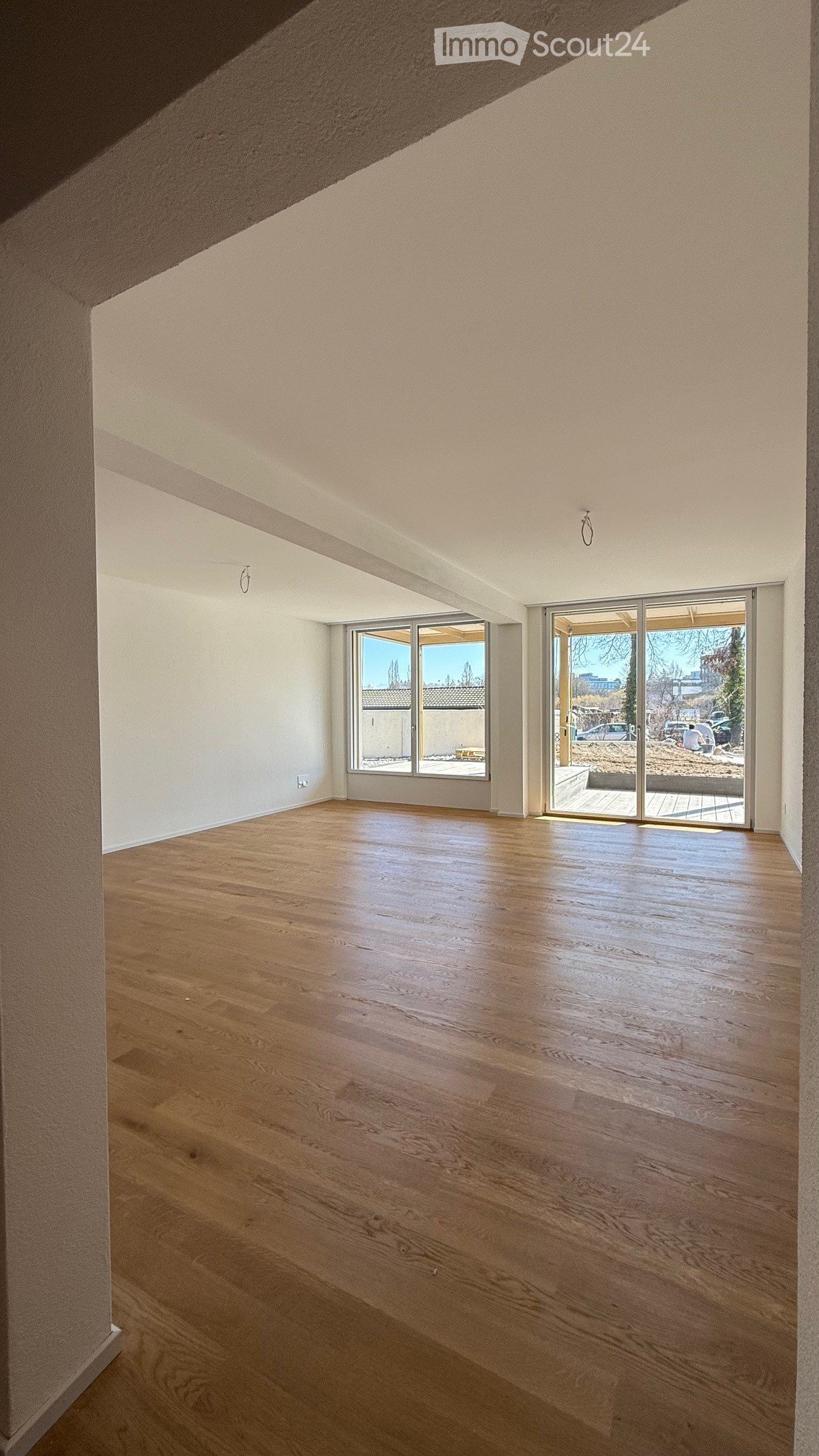 Empty room with wooden flooring, white walls, and large windows and sliding doors leading to an outdoor area.