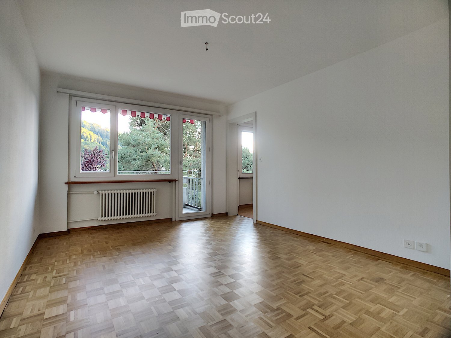 Empty room with wooden herringbone flooring, white walls, large windows with red awnings, radiator, and a door leading to a balcony.