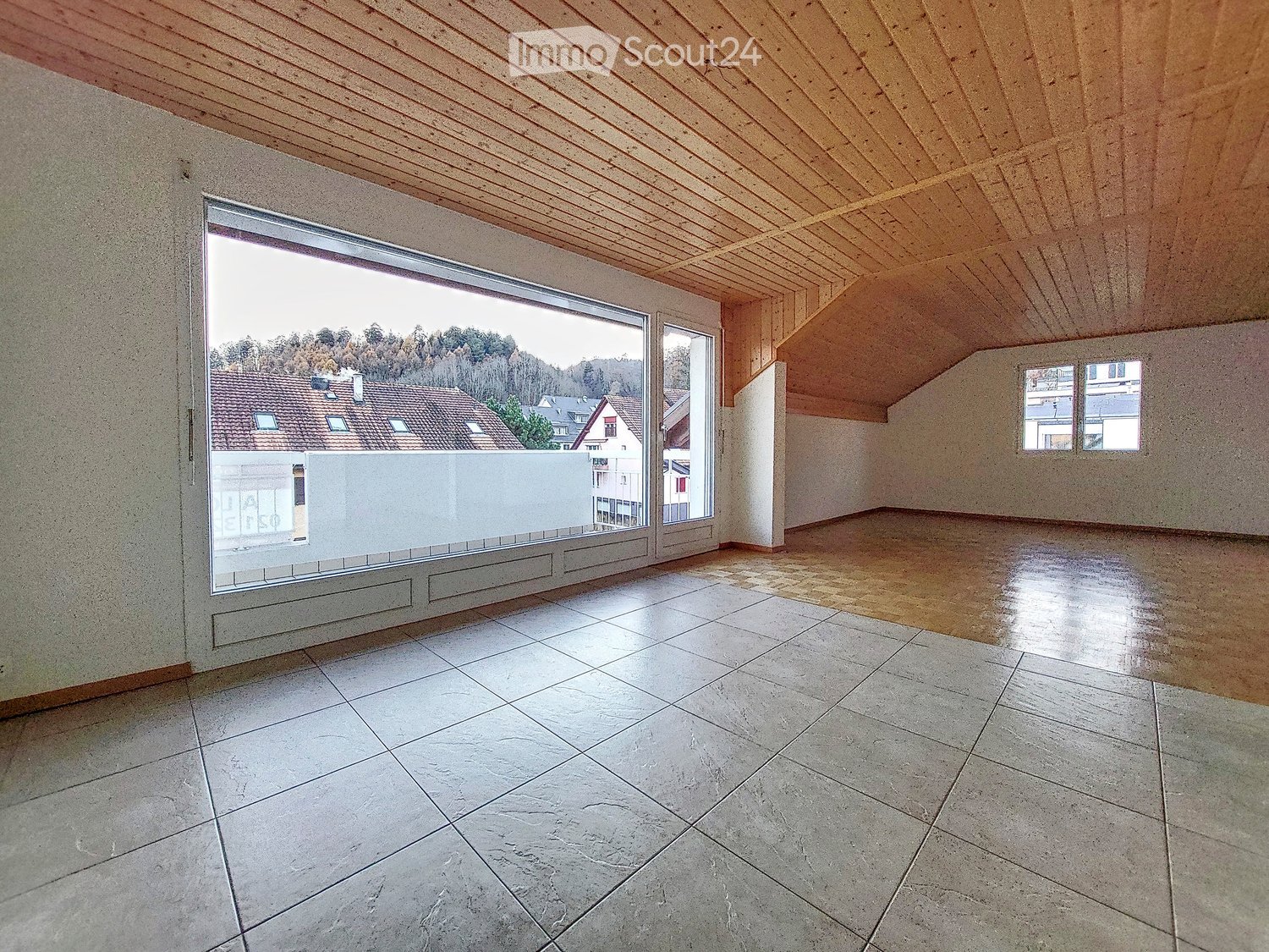Empty room with tiled floor, wooden slanted ceiling, large window offering a view of the neighborhood, and small windows on the side walls.
