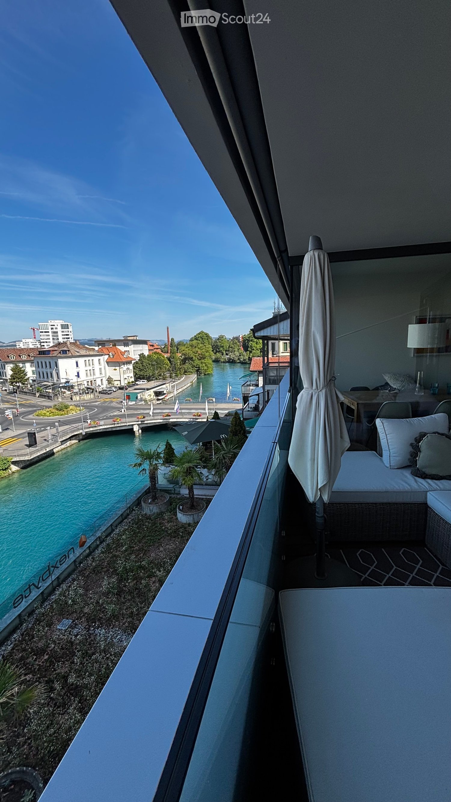 Balcony with a view of a river, featuring a sofa, table, chairs, and an umbrella.