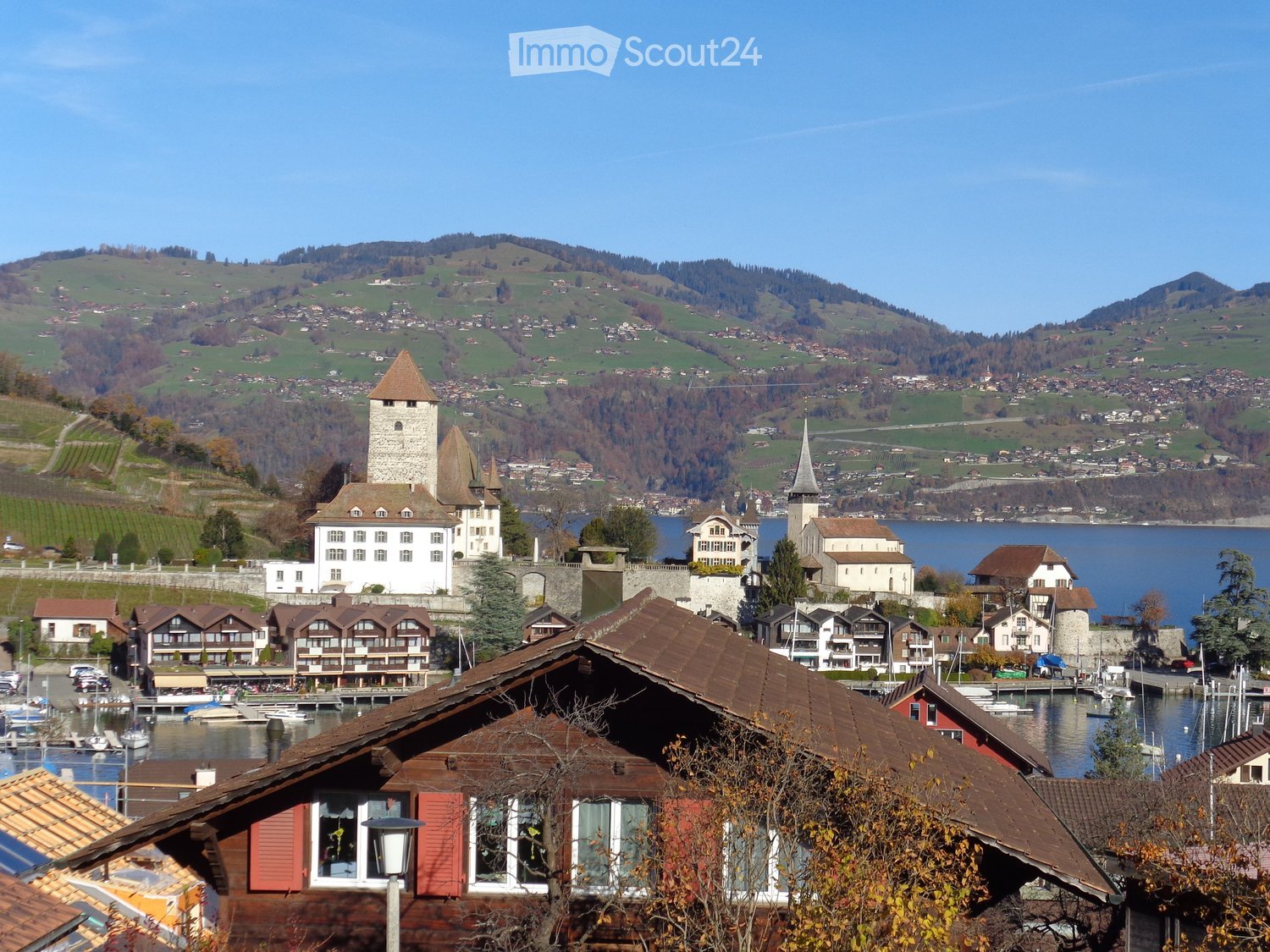 Scenic view of a town with historic buildings, a castle, and a lake surrounded by green hills and mountains.