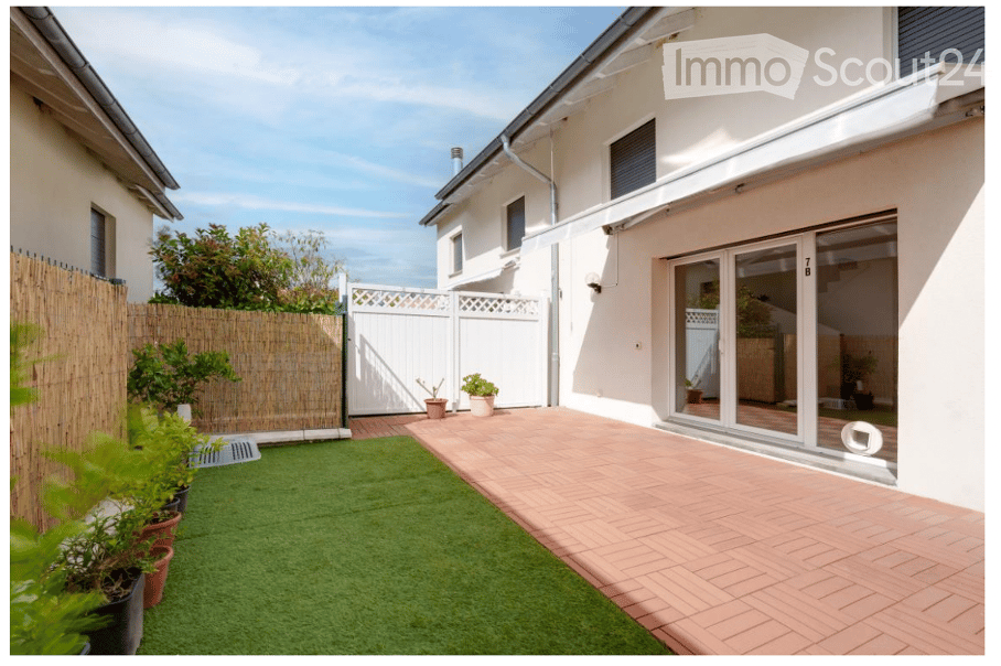 A well-maintained garden area with artificial grass, potted plants, and a brick-paved patio adjacent to a modern house with large glass doors.