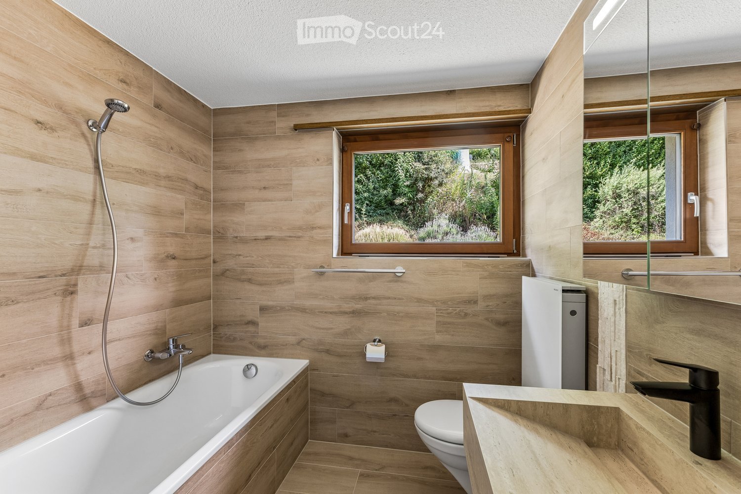 Modern bathroom with wooden paneling, a bathtub, toilet, sink, and a large mirror. Features a showerhead and a towel rack.