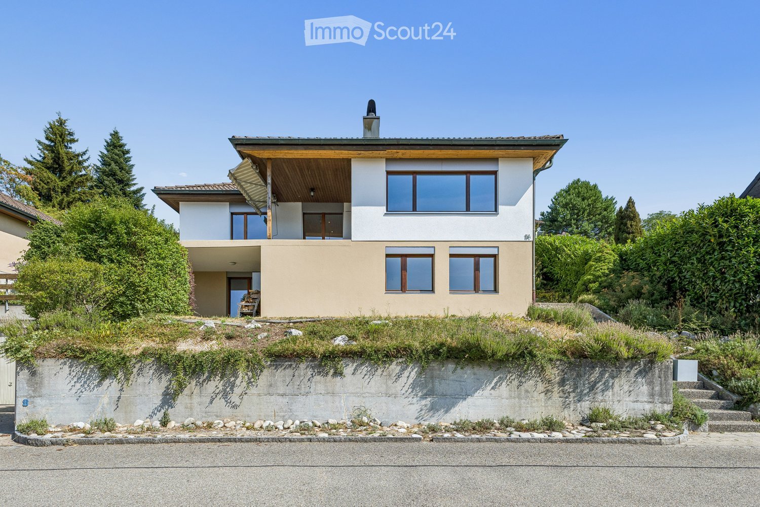 Modern two-story house with a combination of white and beige exterior walls, large windows, and a flat roof with a small chimney. The house features a raised ground floor with a landscaped garden and concrete retaining wall in front.