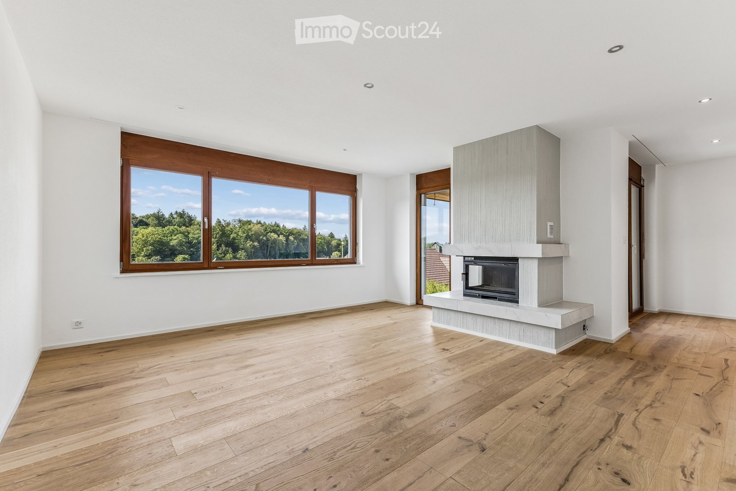 Empty living room with wooden flooring, white walls, large windows, and a built-in fireplace.