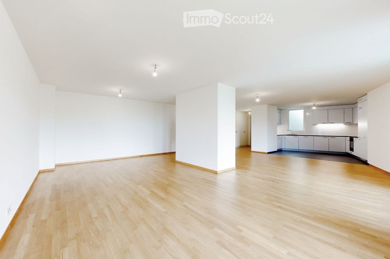 Bright, empty room with light wooden flooring and white walls. A small kitchen area is visible in the background with white cabinets and a countertop.
