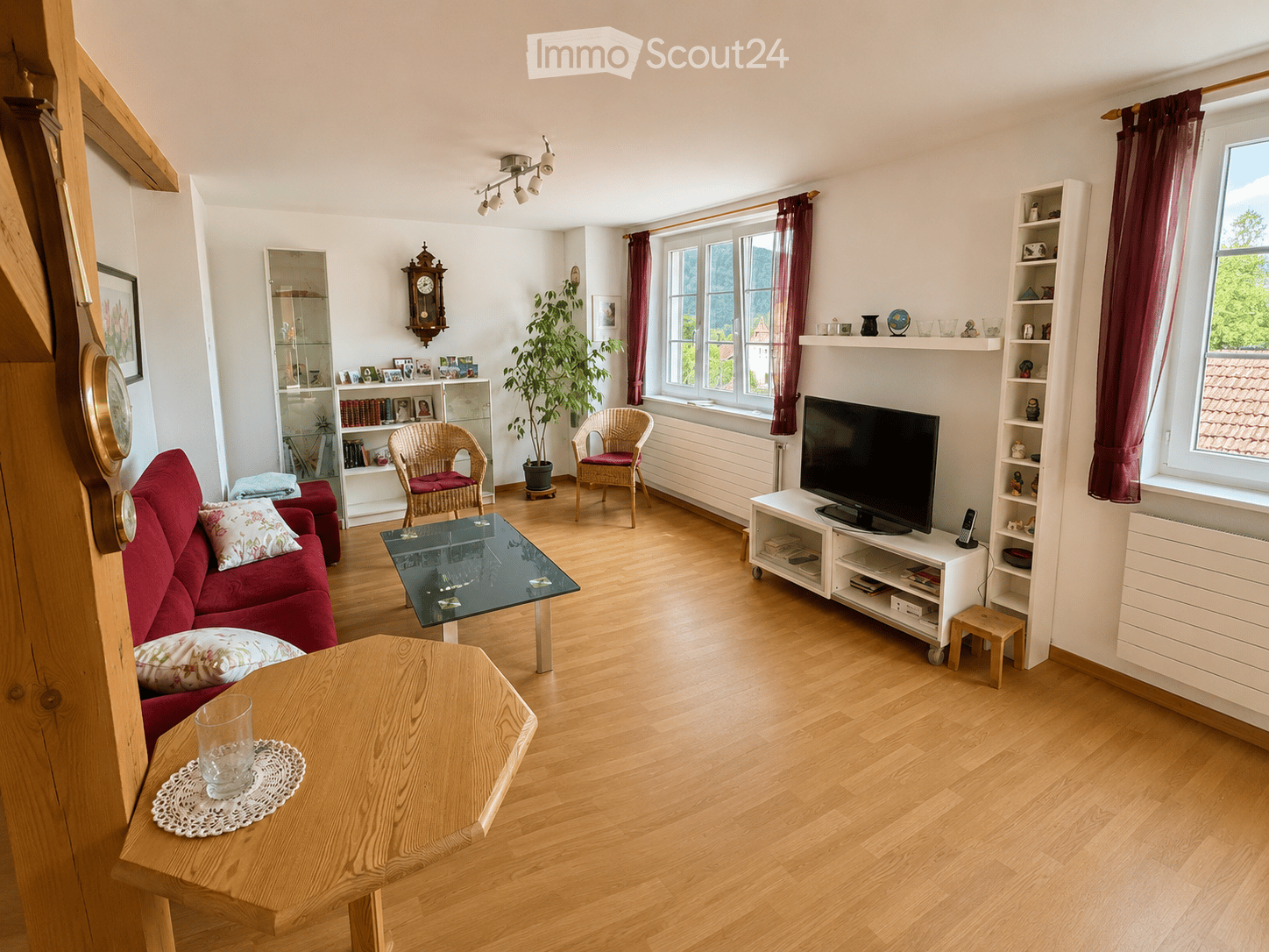 Bright living room with wooden flooring, large windows, red sofa, glass coffee table, wicker chairs, TV stand, and decorative shelves.