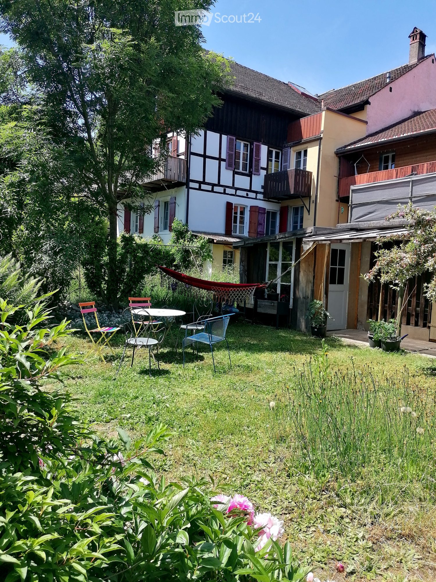 Garden area with a table, chairs, hammock, and surrounding greenery near a multi-story house.