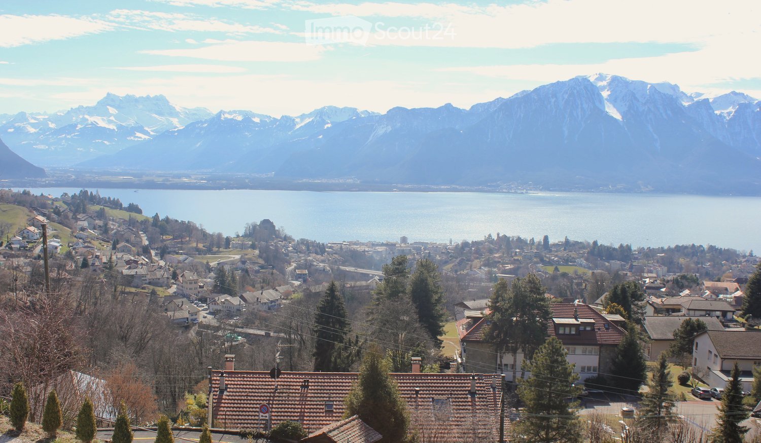 A panoramic view of a town with houses, trees, and a lake surrounded by snow-capped mountains in the background.