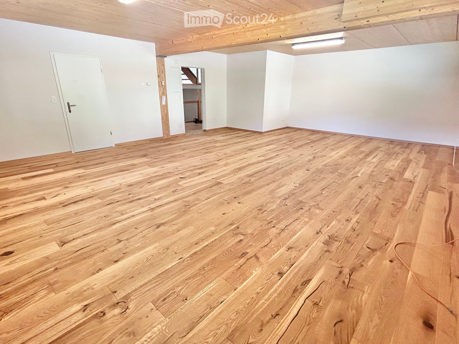 Empty room with wooden flooring, white walls, and exposed wooden beams on the ceiling. Features a white door and a visible staircase leading to another level.