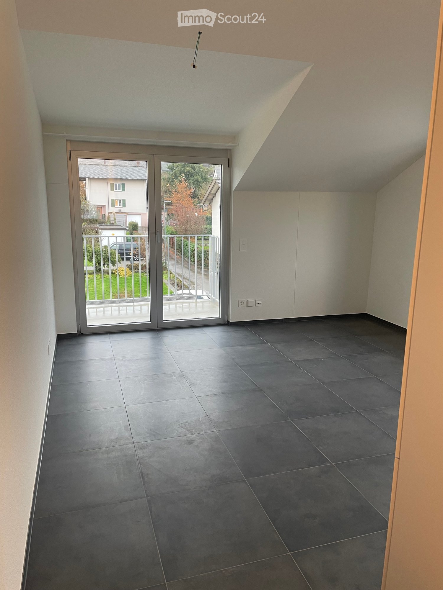 Empty room with dark grey tiled floor, white walls, and a sliding glass door leading to a balcony with a metal railing.