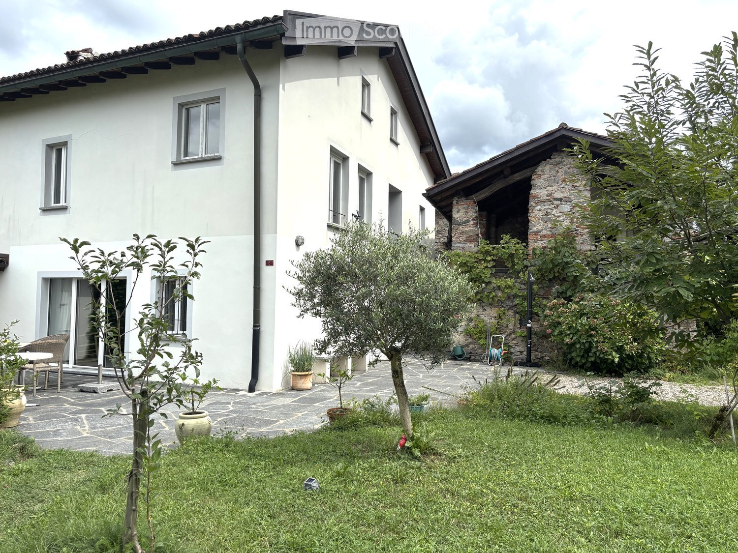 Two-story white house with multiple windows, a stone chimney, and a well-maintained garden with olive trees, potted plants, and a patio area.