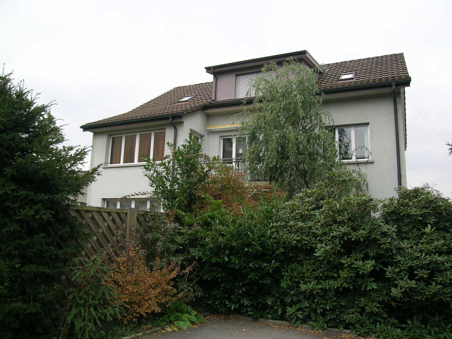 2 story house, white exterior, brown roof, balcony, large windows, surrounded by greenery