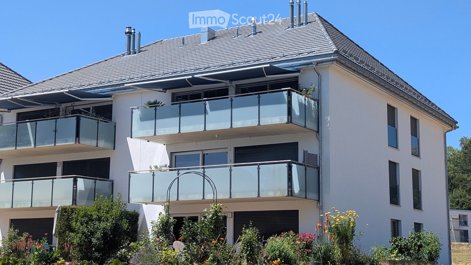Modern two-story house with white exterior, grey roof, multiple balconies with glass railings, and well-maintained garden.
