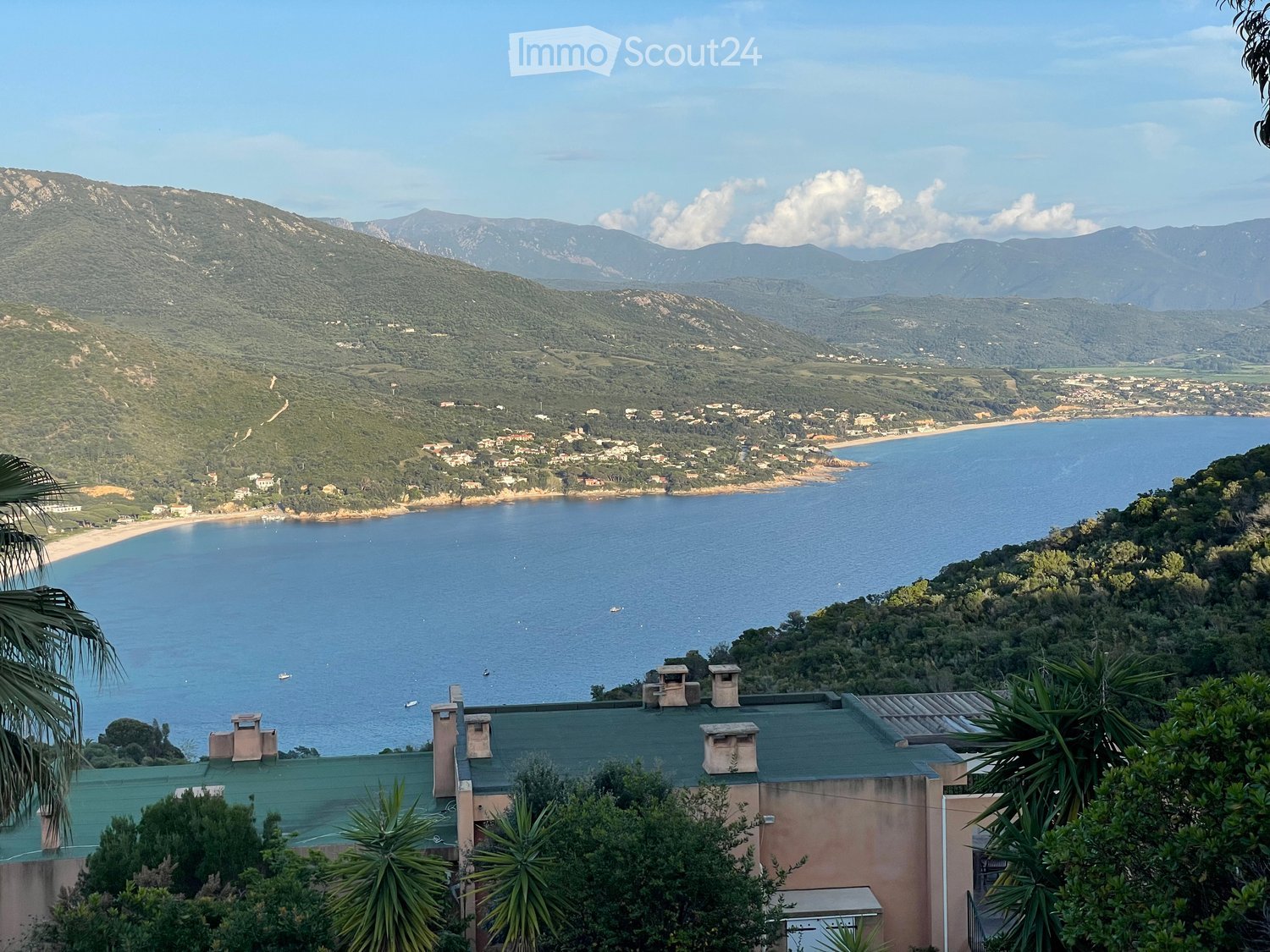 A scenic coastal view with a house on a hillside overlooking the sea, surrounded by lush greenery and distant mountains.
