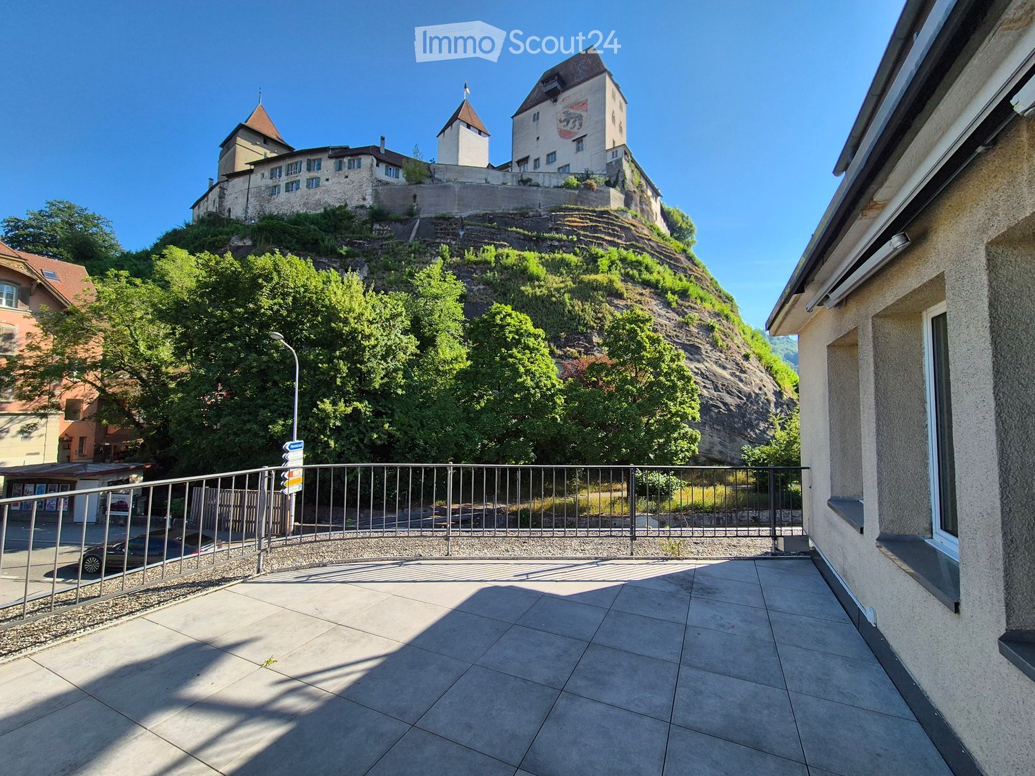 Balcony with tiled floor, metal railing, overlooking a historic castle on a hill, surrounded by trees and greenery.