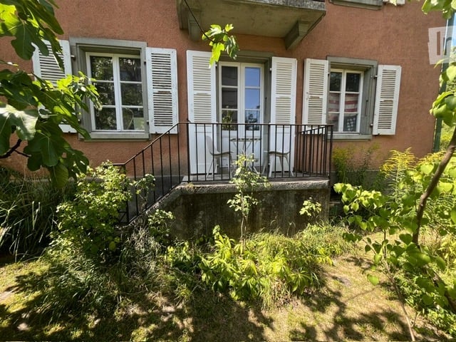 Two-story building with a balcony, white shutters, and a small garden area.