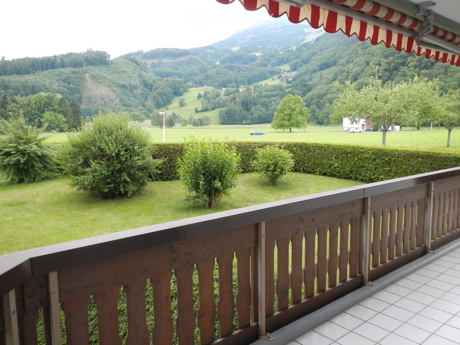 Balcony with wooden railing, tiled floor, and scenic view of green fields, trees, and distant mountains.