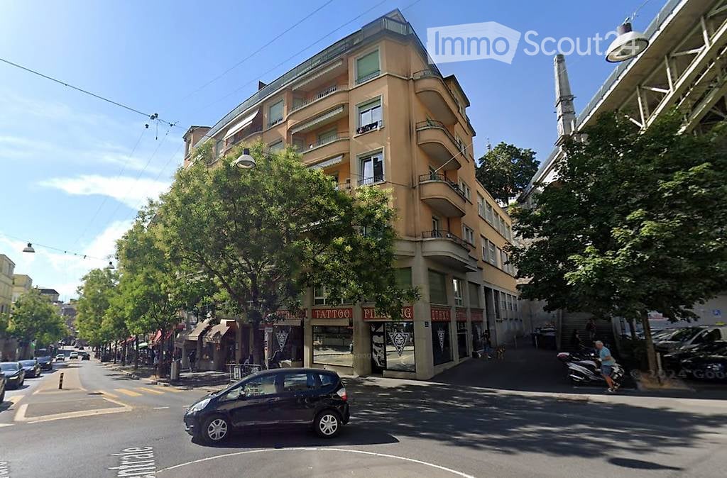 Multi-story building with balconies and storefronts on the ground floor, located on a tree-lined street with parked cars and pedestrians.