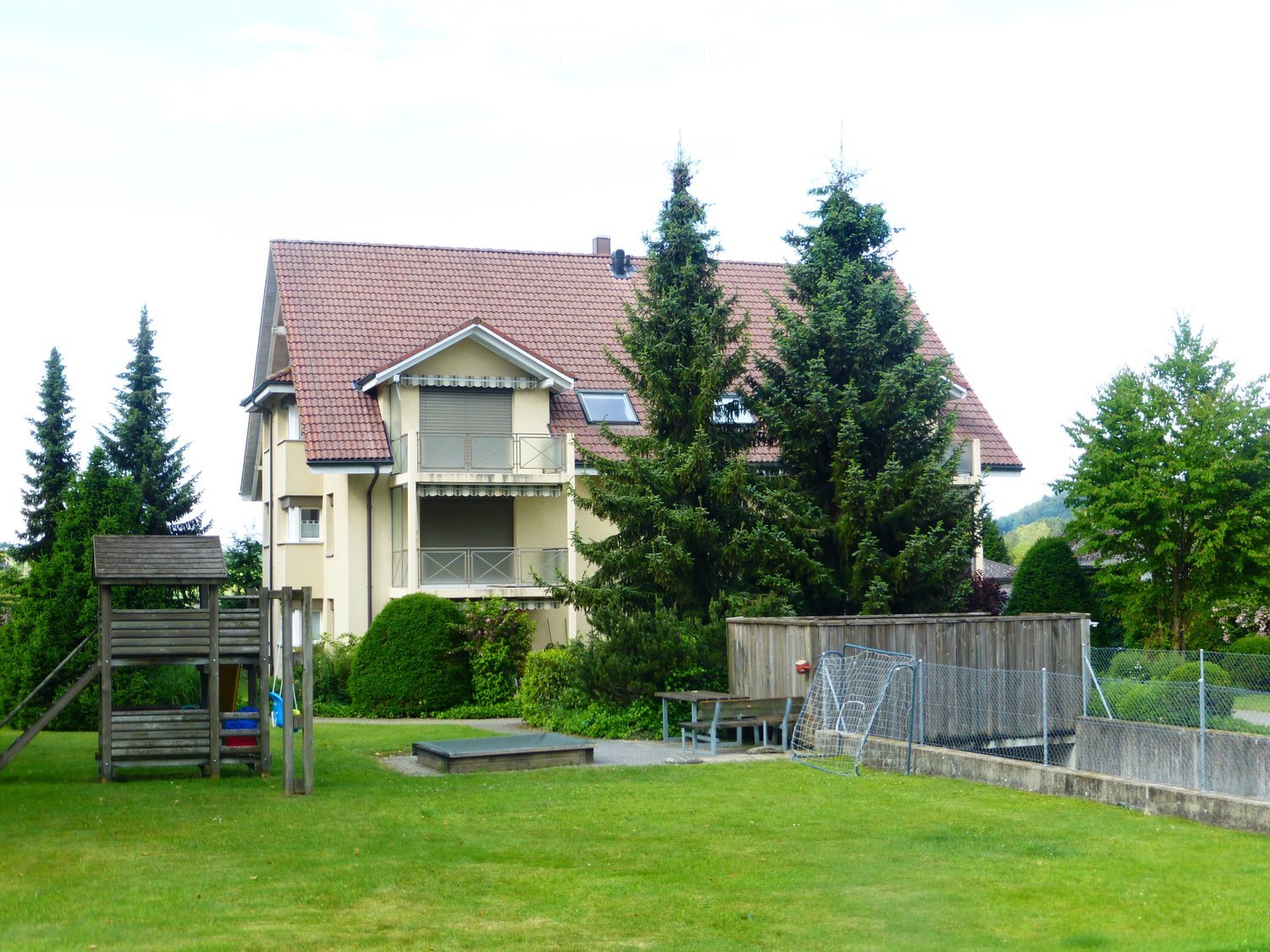 Two-story house with red-tiled roof, balcony, surrounded by trees and a well-maintained lawn with playground equipment.