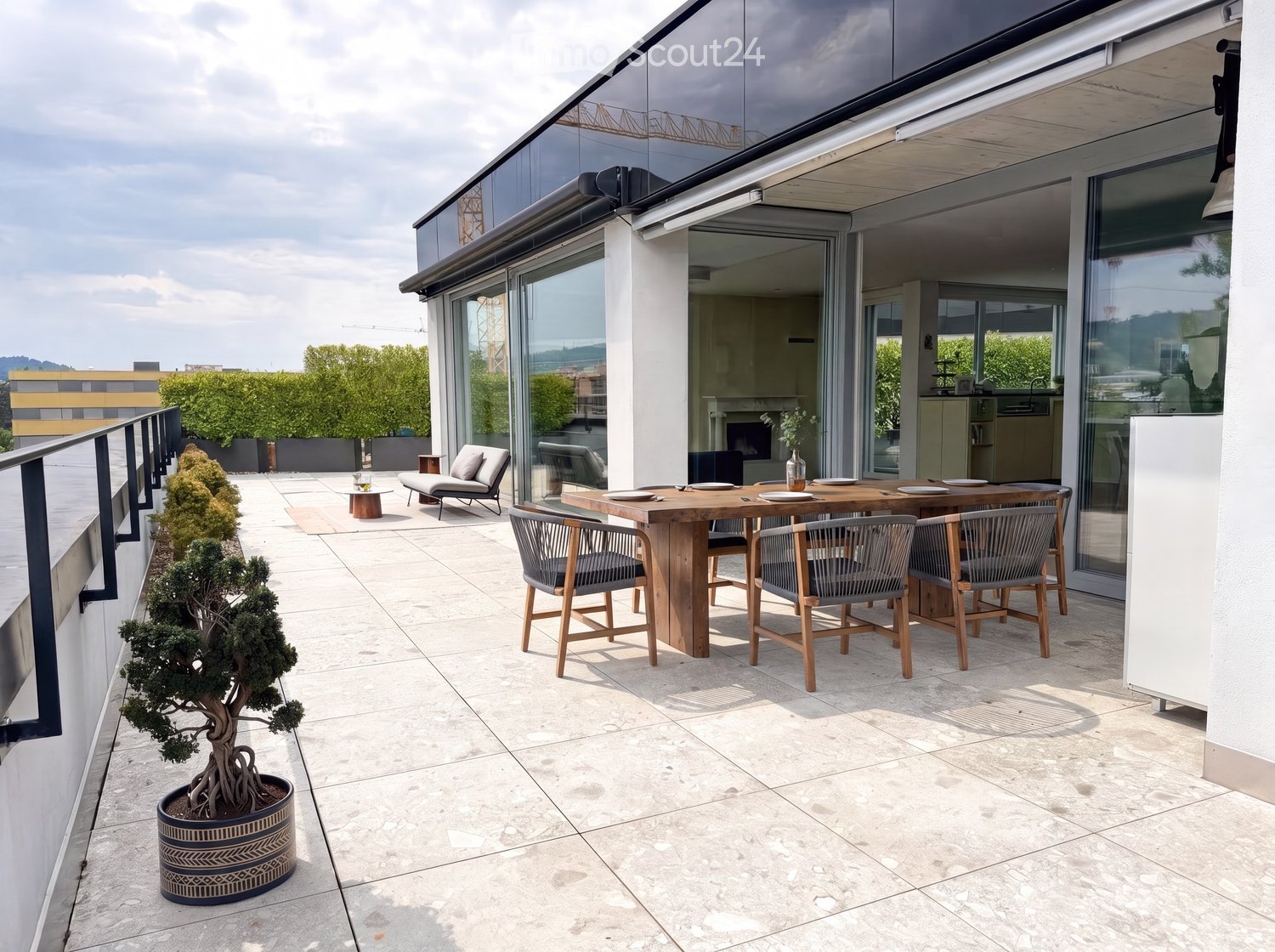 Modern rooftop terrace with a wooden dining table, chairs, a lounge chair, and a small tree in a pot.