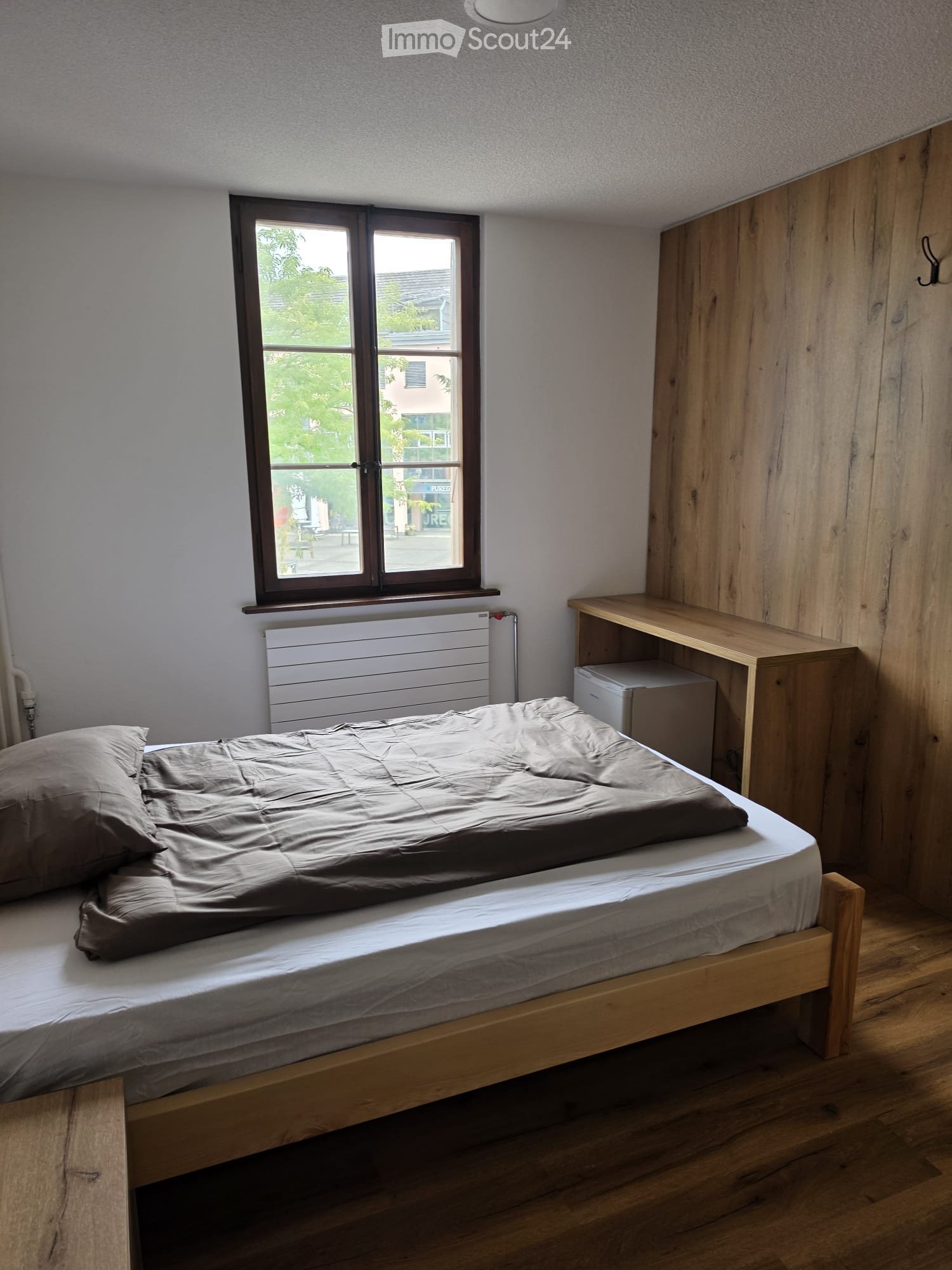 Cozy bedroom with a wooden bed, grey bedding, a radiator below a window, a wooden wardrobe, and a small shelf.