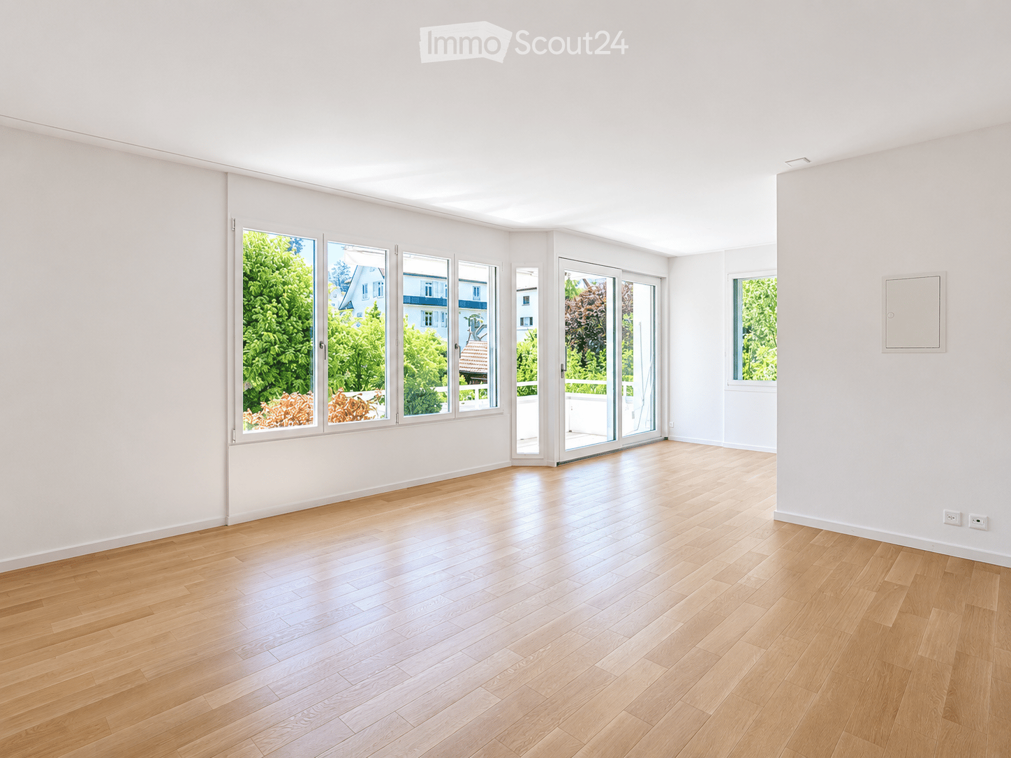 Empty room with light wooden flooring and white walls, featuring large windows and a sliding glass door leading to a balcony.