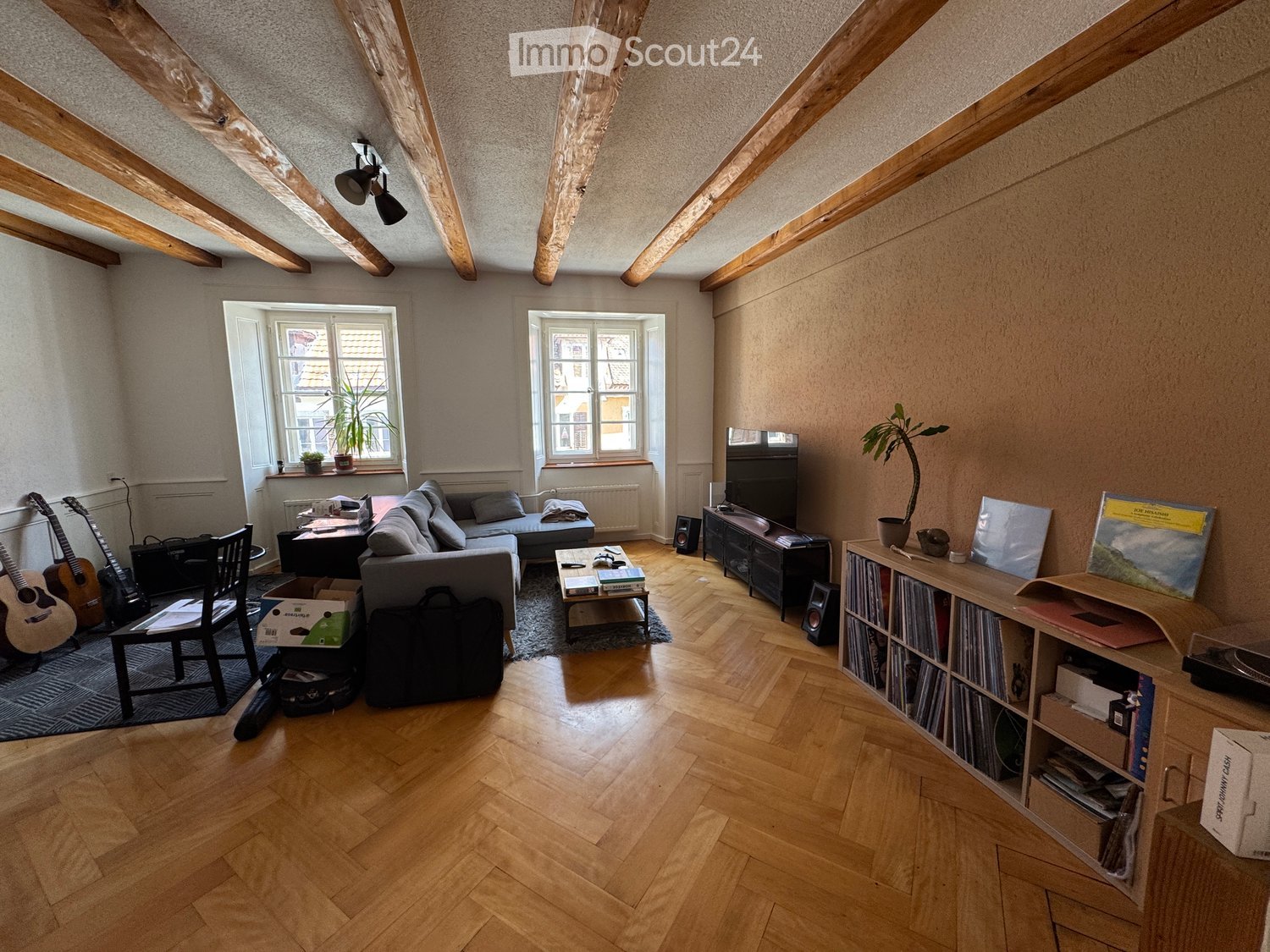 Living room with wooden herringbone flooring, exposed wooden beams, large windows, grey sofa, coffee table, guitar, bookshelf, and various decorative items.
