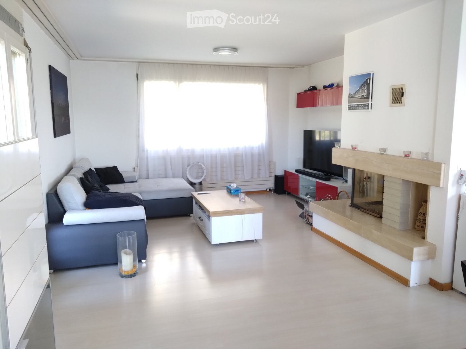 Modern living room with white walls, grey sofa, coffee table, fireplace, and TV. Features large windows with white curtains and a sleek design.
