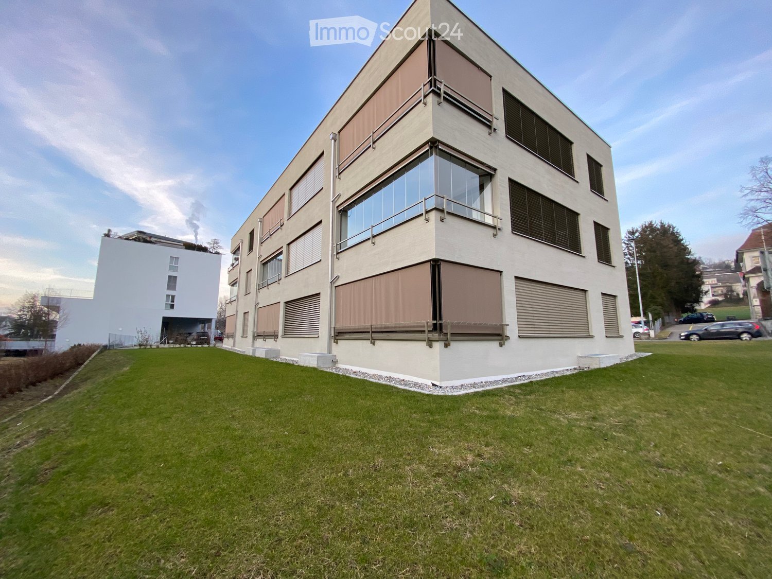 Modern apartment building with glass windows, beige exterior, and a grassy lawn in front.