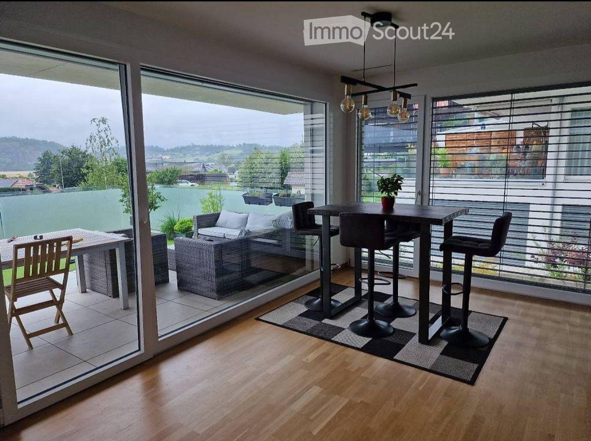 Modern living room with large windows offering a scenic view, featuring a sofa, coffee table, bar stools, a chair, and a small potted plant on the table.