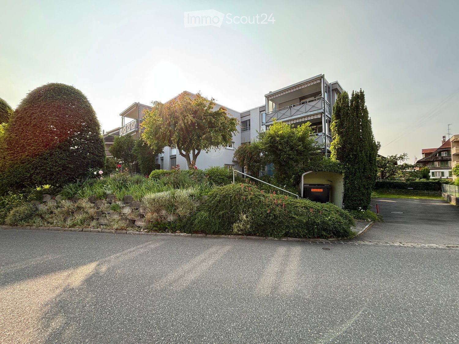 Modern multi-story residential building with balconies, surrounded by well-maintained gardens and trees.