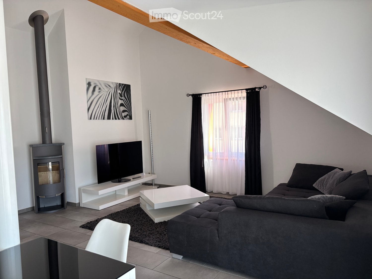 Modern living room with a dark grey sectional sofa, white coffee table, flat-screen TV on a white stand, fireplace, and large window with curtains.
