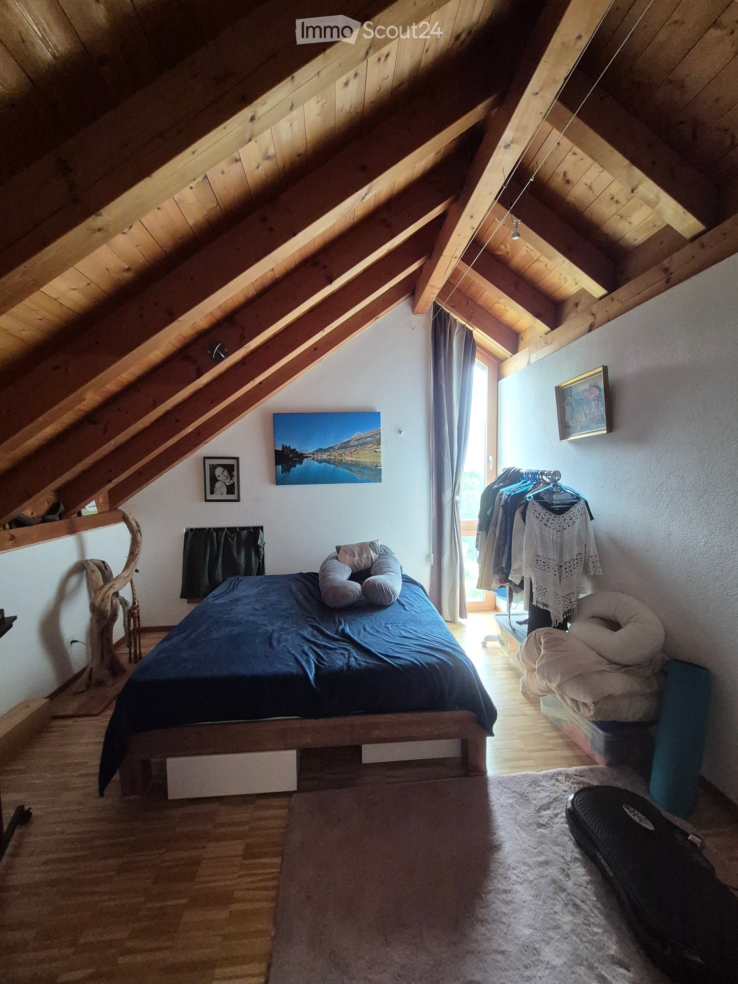 Cozy attic bedroom with wooden slanted ceilings, a large bed with dark blue bedding, pillows, a mirror, a clothing rack, and artwork on the walls.