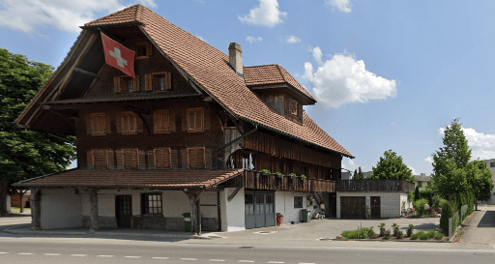 Traditional Swiss wooden house with a red-tiled roof, multiple stories, and a Swiss flag. The building features large windows and a covered porch area.